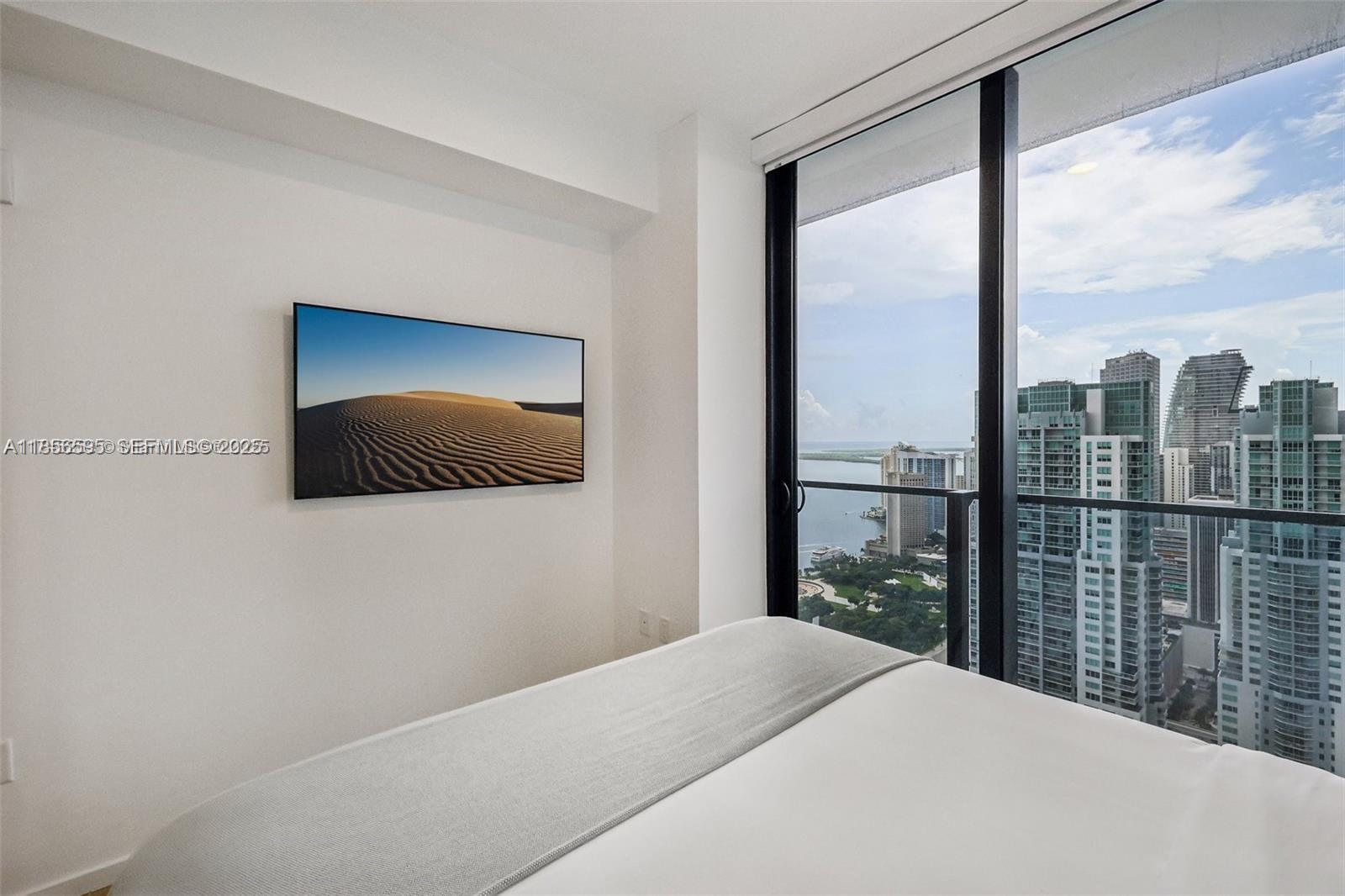 A bedroom features a wall-mounted television, white bedding, and floor-to-ceiling windows overlooking a city skyline — 398 NE 5th St # 4415, Miami FL 33132, MIAMI, FL