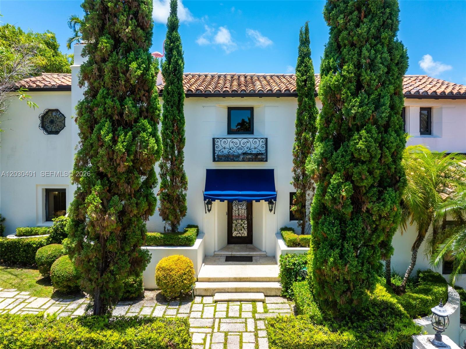 A white stucco house features a blue awning over the front door and tall cypress trees — 5860 Pine Tree Dr, Miami Beach FL 33140, MIAMI BEACH, FL