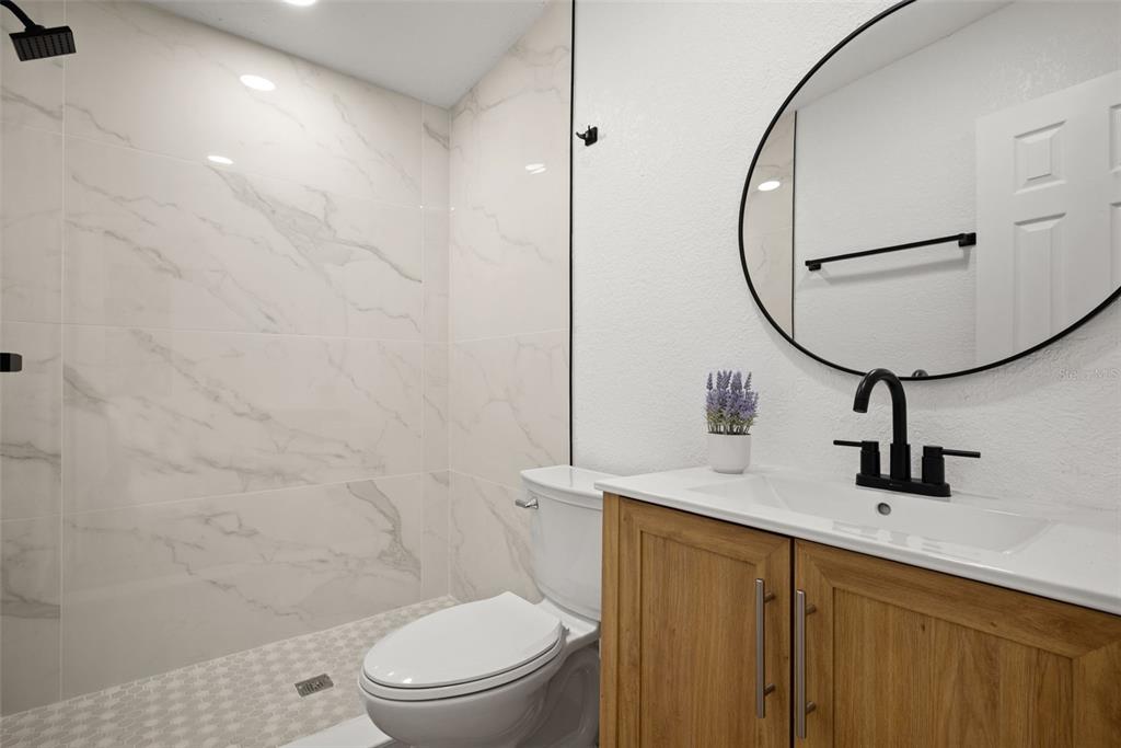 A bathroom features a walk-in shower with marble-look tile, a vanity, and a round mirror — 9596 GROVELAND STREET, SEMINOLE, FL
