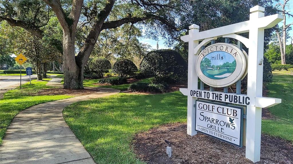 A white wooden sign for Victoria Hills Golf Club stands beside a paved walkway — 171 BIRCHMONT DRIVE, DELAND, FL