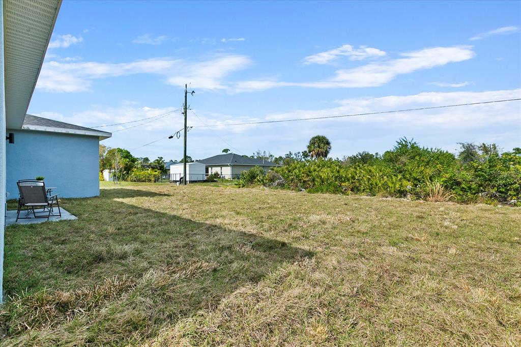 A grassy backyard extends from a blue house with a small patio and chair — 18400 TULANE AVENUE, PORT CHARLOTTE, FL
