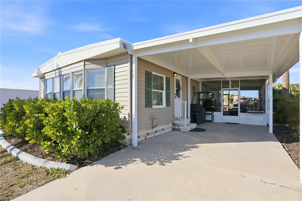 A tan manufactured home features a large carport, a screened-in porch, and a landscaped front yard — 1427 SANDERLING DRIVE, ENGLEWOOD, FL