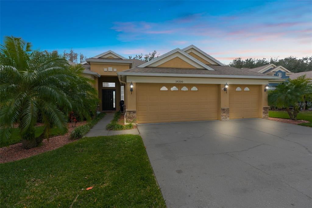 A tan single-story house with a three-car garage and a palm tree in the front yard — 19200 SUNSET BAY DRIVE, LAND O LAKES, FL
