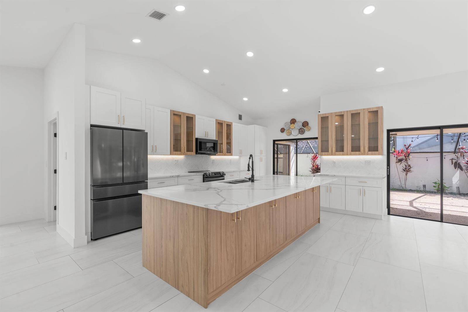 A modern kitchen features white cabinets, wood-toned accents, a large marble island, and sliding doors — 21326 Campo Allegro Drive, Boca Raton FL 33433, BOCA RATON, FL