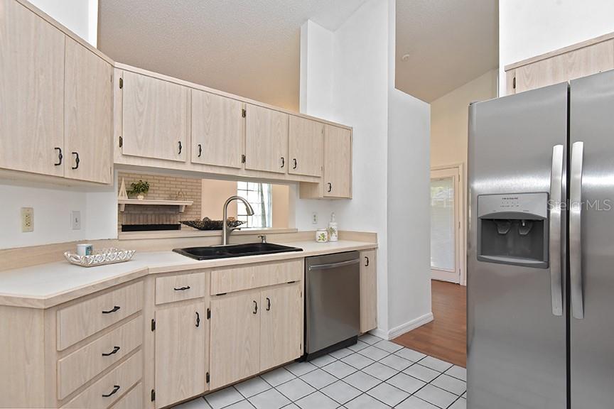A kitchen features light wood cabinets, stainless steel appliances, and a pass-through window to the living area — 2213 PINE TREE DRIVE, EDGEWATER, FL