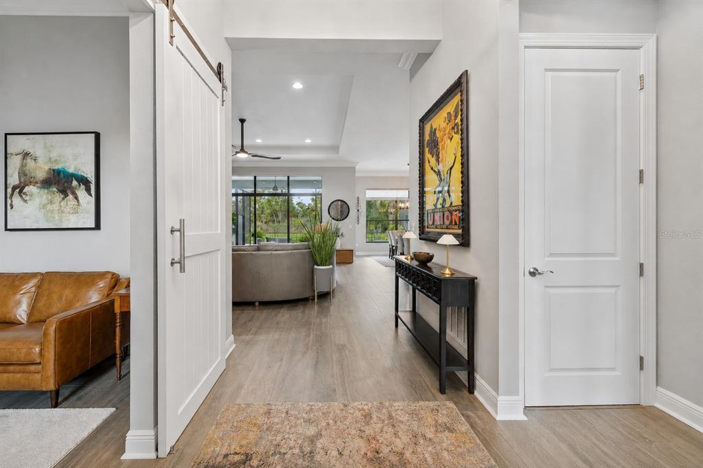 A hallway with light wood flooring leads into a living room with a sliding barn door — 9005 BERNINI PLACE, SARASOTA, FL
