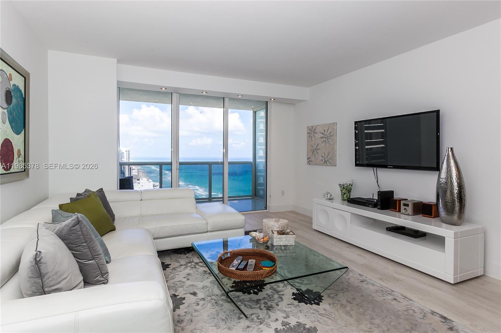 A modern living room features a white sectional sofa, glass coffee table, and ocean views — 1830 S OCEAN DR. # 3711, Hallandale Beach FL 33009, HALLANDALE BEACH, FL
