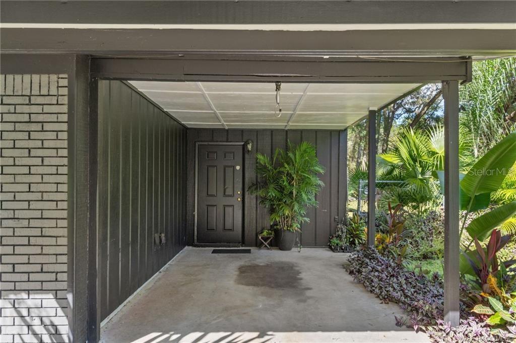 A covered concrete entryway features a dark-painted door, vertical siding, and surrounding tropical landscaping — 3751 SE 145TH STREET, SUMMERFIELD, FL