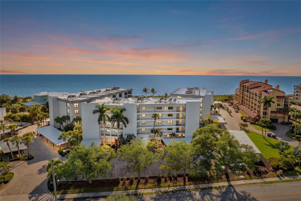 An aerial view shows a multi-story white apartment building near the ocean at sunset — 8764 MIDNIGHT PASS ROAD UNIT A201, SARASOTA, FL