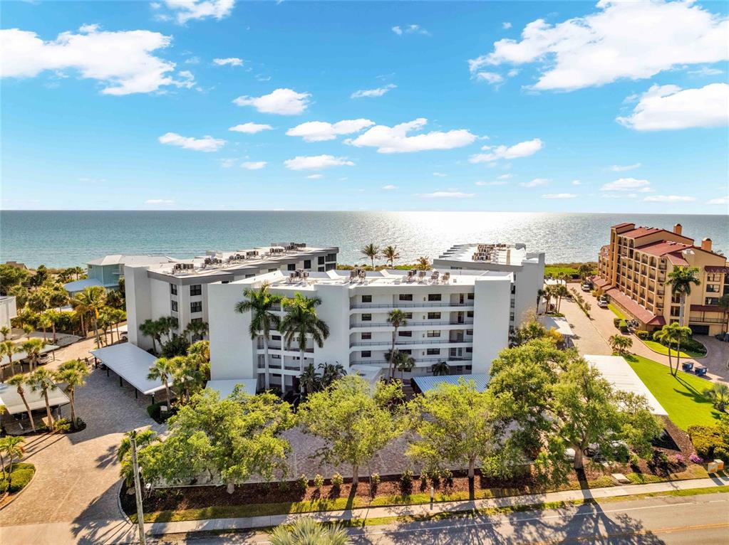 An aerial view shows a multi-story white apartment building near the ocean with palm trees — 8764 MIDNIGHT PASS ROAD UNIT A201, SARASOTA, FL