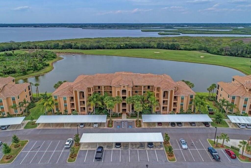 An aerial view shows a multi-story apartment building with a parking lot and a lake — 6515 GRAND ESTUARY TRAIL UNIT 403, BRADENTON, FL