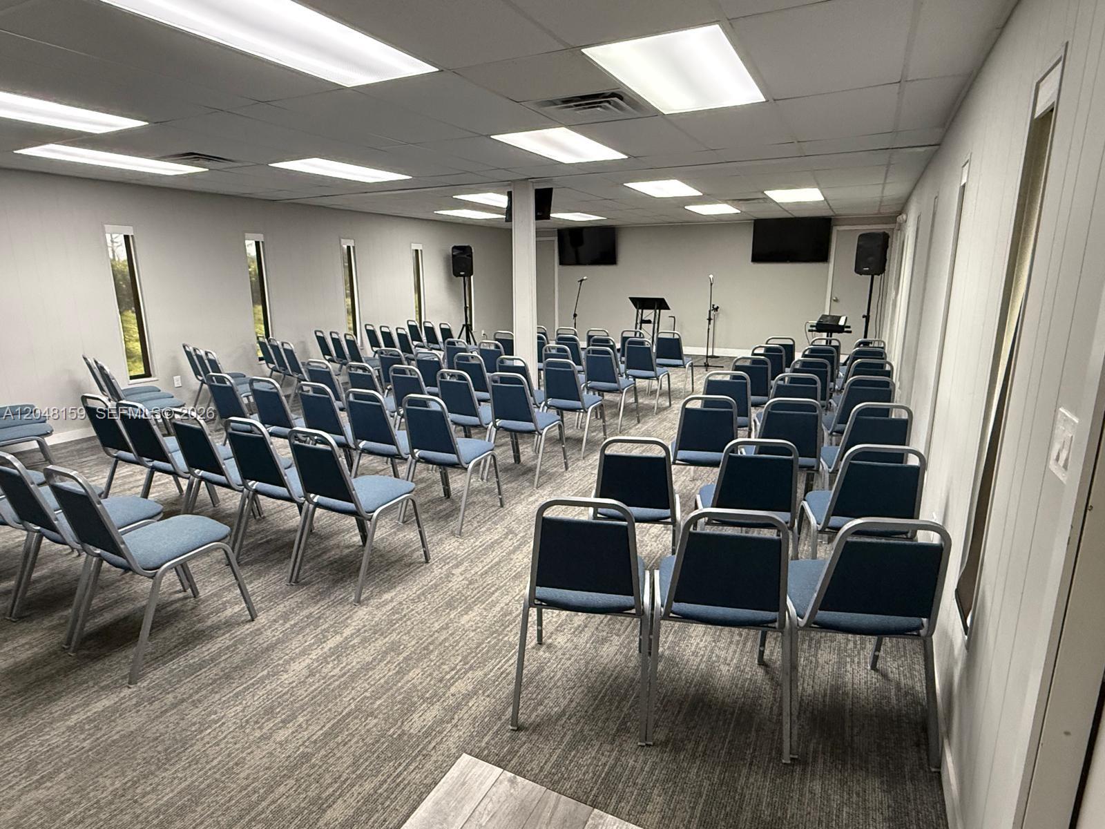 A meeting room features rows of blue chairs, gray carpeting, and a small stage area — 18500 Griffin Rd, Southwest Ranches FL 33332, SOUTHWEST RANCHES, FL