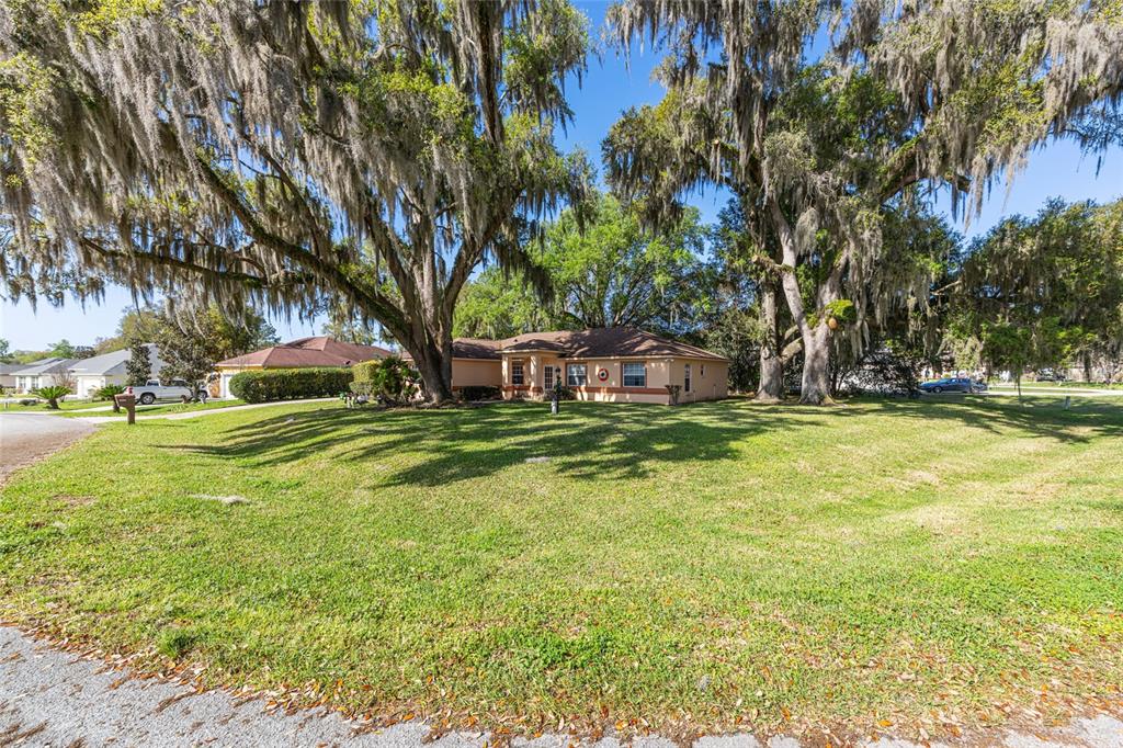 A single-story house with a tan exterior sits on a grassy lot under large trees — 10090 SW 78TH COURT, OCALA, FL