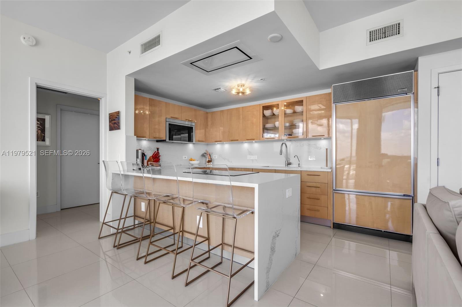A modern kitchen features light wood cabinets, a white marble island, and clear bar stools — 6899 Collins Ave # 2403, Miami Beach FL 33141, MIAMI BEACH, FL