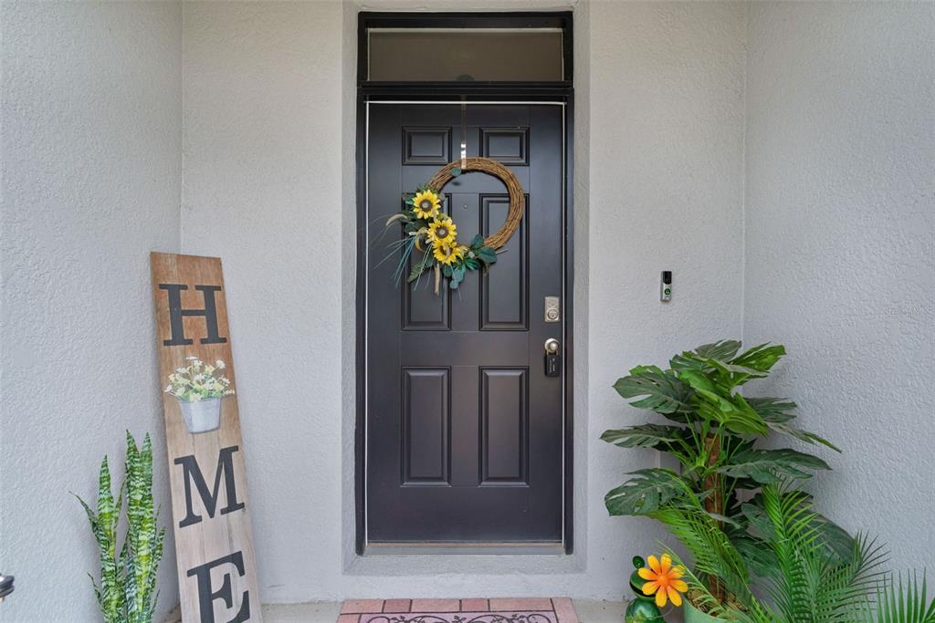 A dark front door with a sunflower wreath sits between a wooden sign and plants — 5365 PINECONE COURT, WILDWOOD, FL