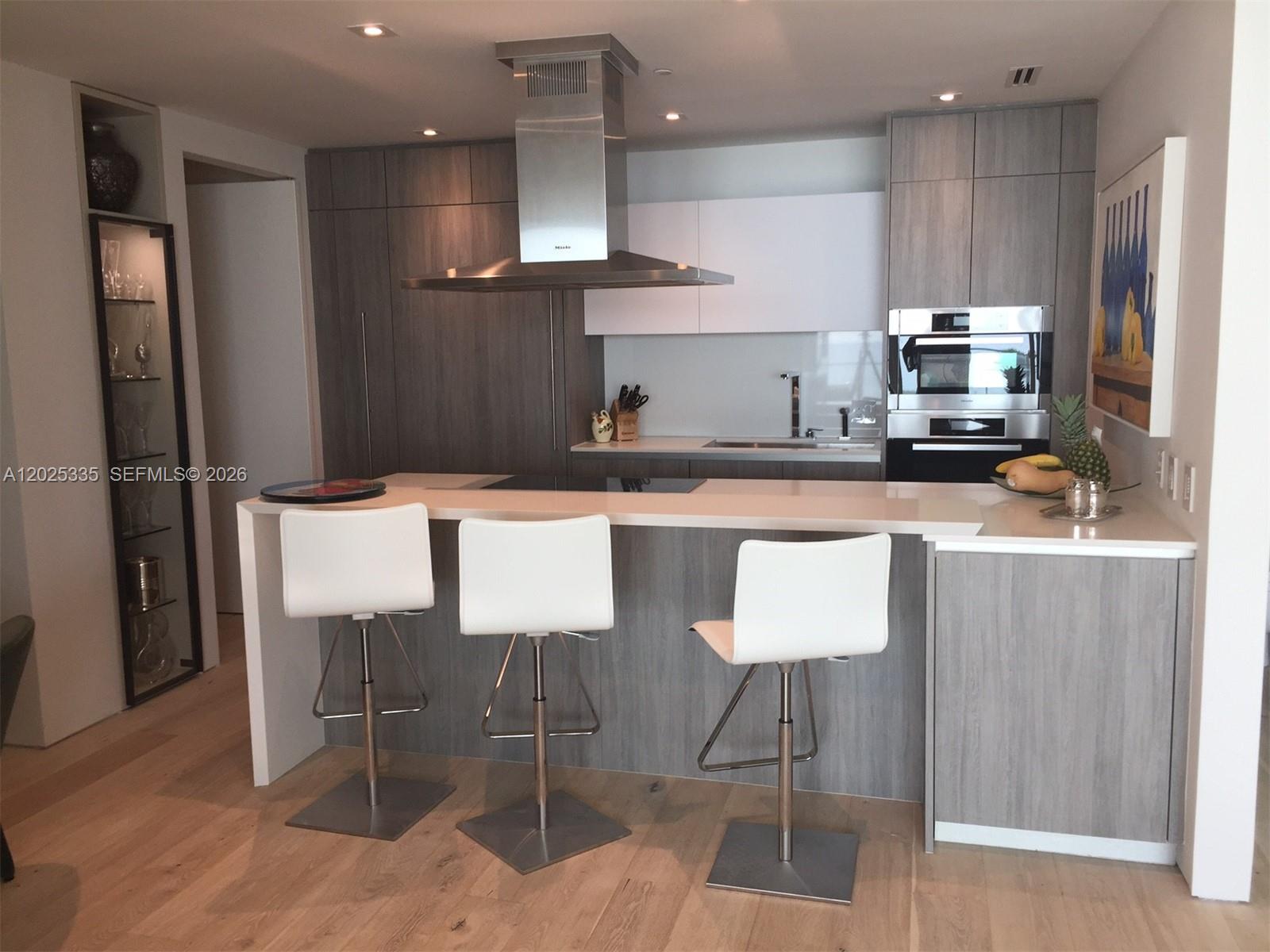 A modern kitchen features gray wood cabinetry, white countertops, three bar stools, and stainless appliances — 350 OCEAN DR # 304N, Key Biscayne FL 33149, KEY BISCAYNE, FL