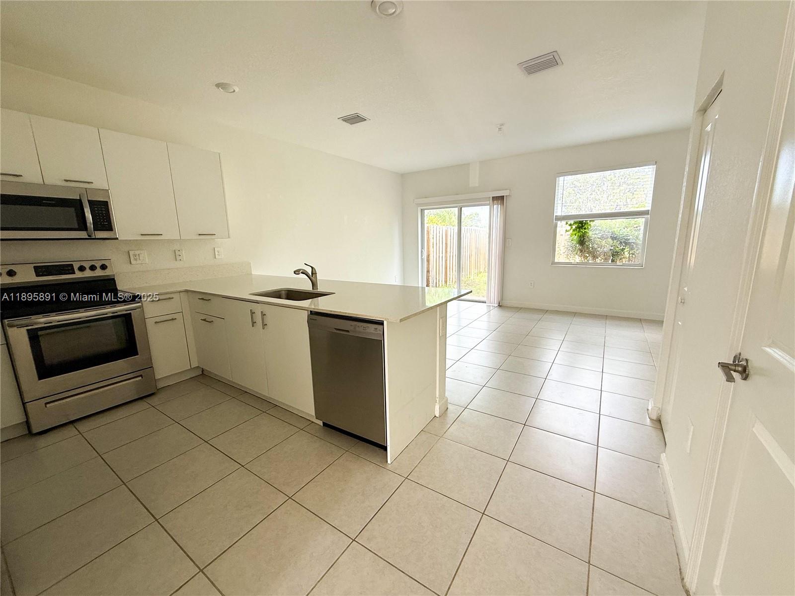 A kitchen with white cabinets, stainless steel appliances, and tile flooring opens to a living area — 12445 NW 23rd Ct, Miami FL 33167, MIAMI, FL
