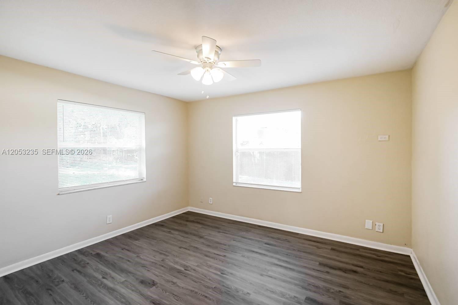 An empty bedroom features light-colored walls, dark wood-look flooring, two windows, and a ceiling fan — DAVIE, FL