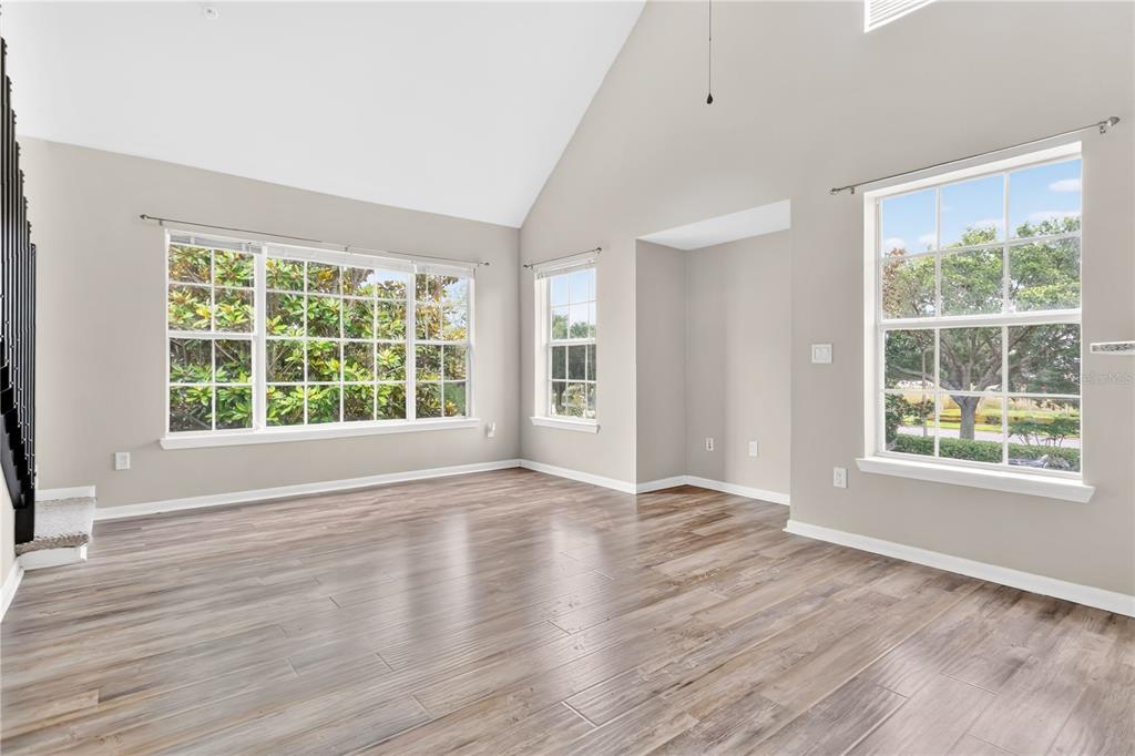 An empty living room with vaulted ceilings, wood-look tile flooring, and multiple large windows — 8925 LEE VISTA BOULEVARD UNIT 2704, ORLANDO, FL