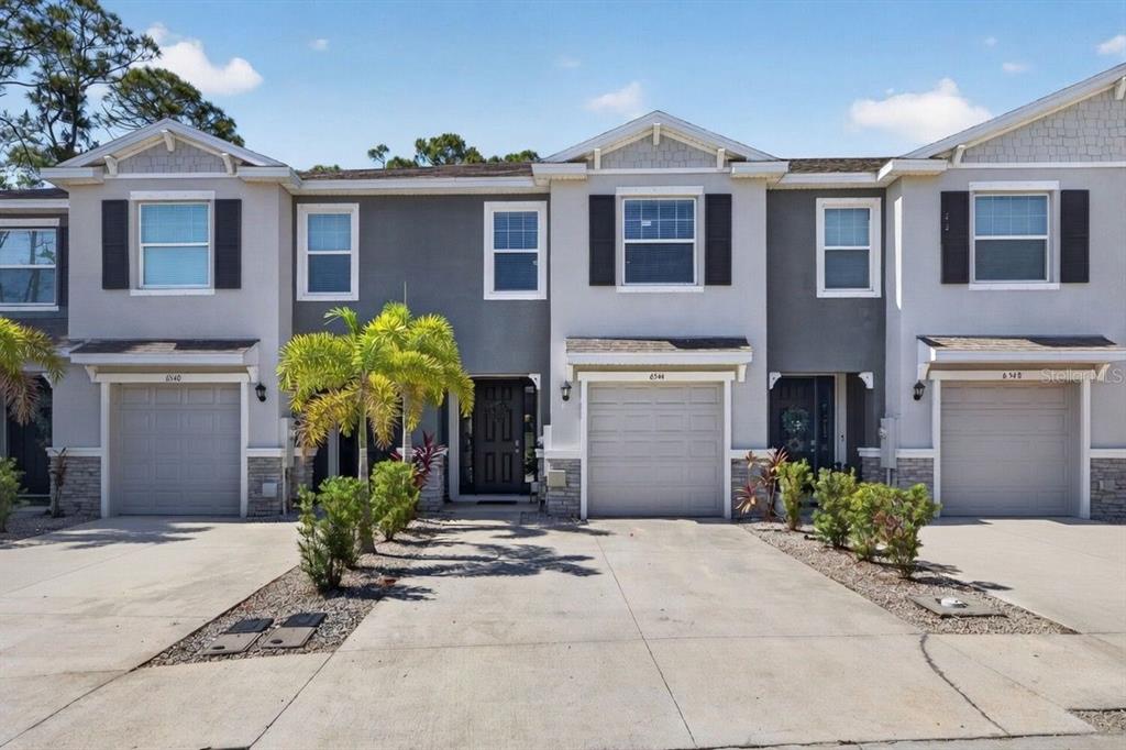 A row of three attached townhomes with gray siding, stone accents, and individual garage doors — 6544 CALYPSO CORAL LANE, SARASOTA, FL