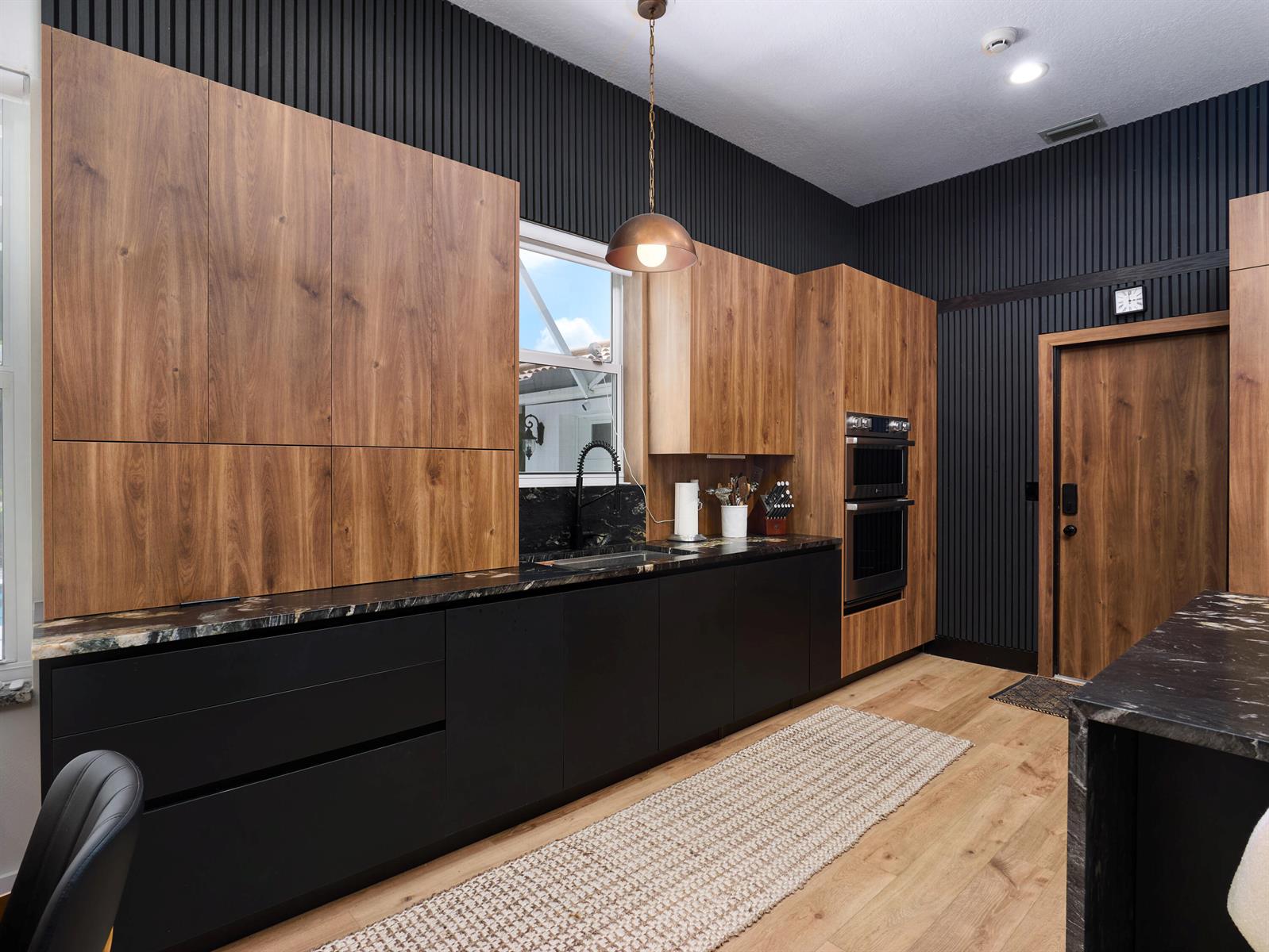 A modern kitchen features black slatted walls, wood-grain cabinetry, black countertops, and a long runner rug — 8166 Quail Meadow Way, West Palm Beach FL 33412, WEST PALM BEACH, FL