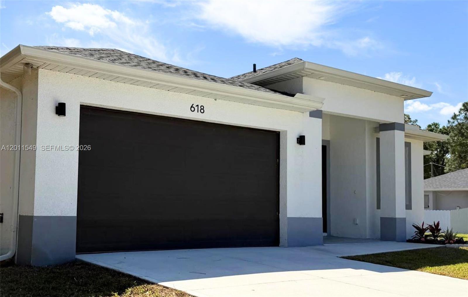 The exterior of a white house features a dark garage door and a covered entryway — 618 SHELDON AVE, Lehigh Acres FL 33936, LEHIGH ACRES, FL