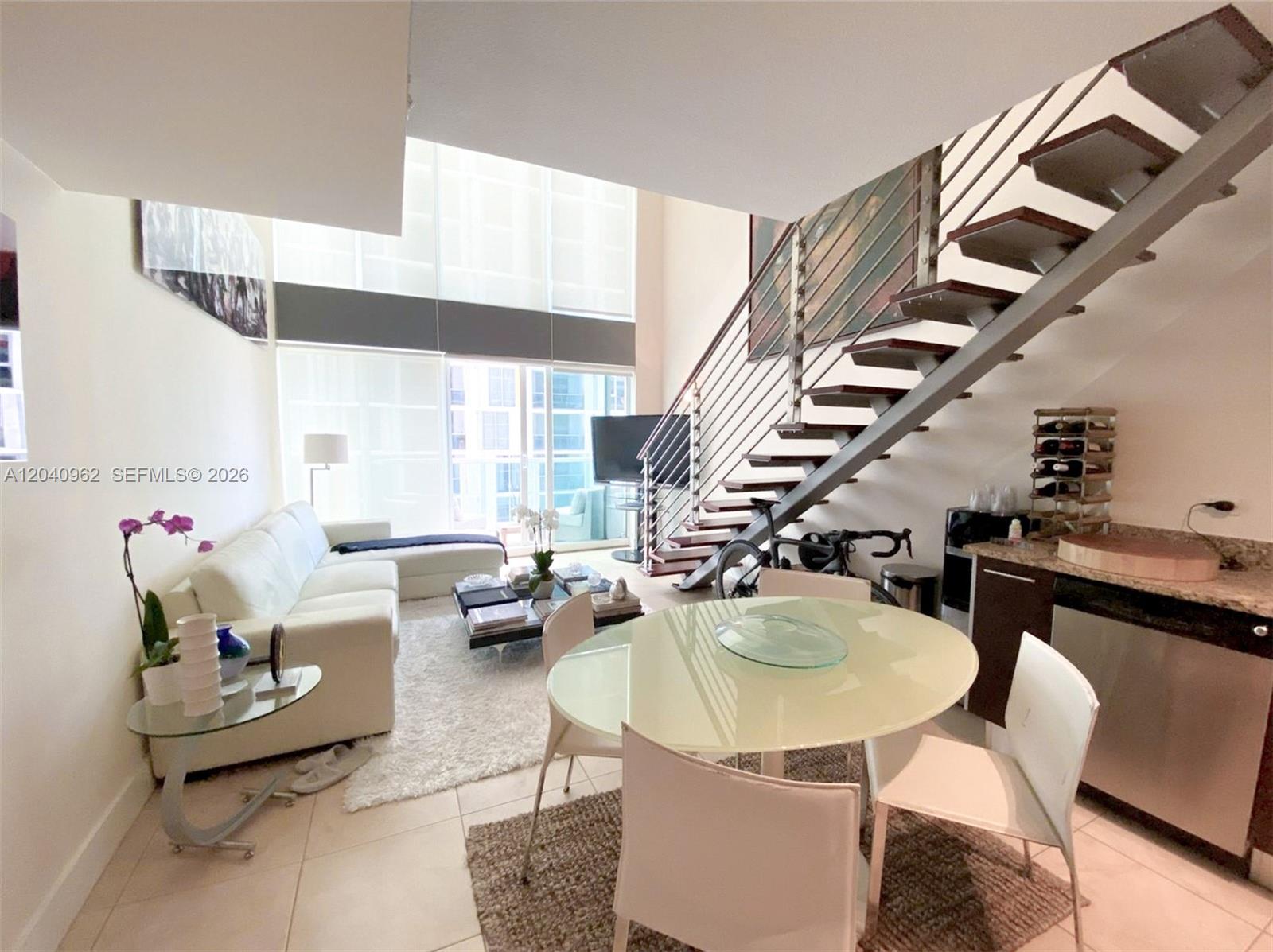 A modern living and dining area features a floating staircase, floor-to-ceiling windows, and tile flooring — 41 SE 5th St # 1006, Miami FL 33131, MIAMI, FL