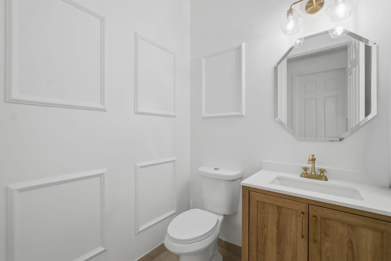A small bathroom features white walls with molding, a wood vanity, and an octagonal mirror — 13769 Moss Agate Avenue, Delray Beach FL 33446, DELRAY BEACH, FL