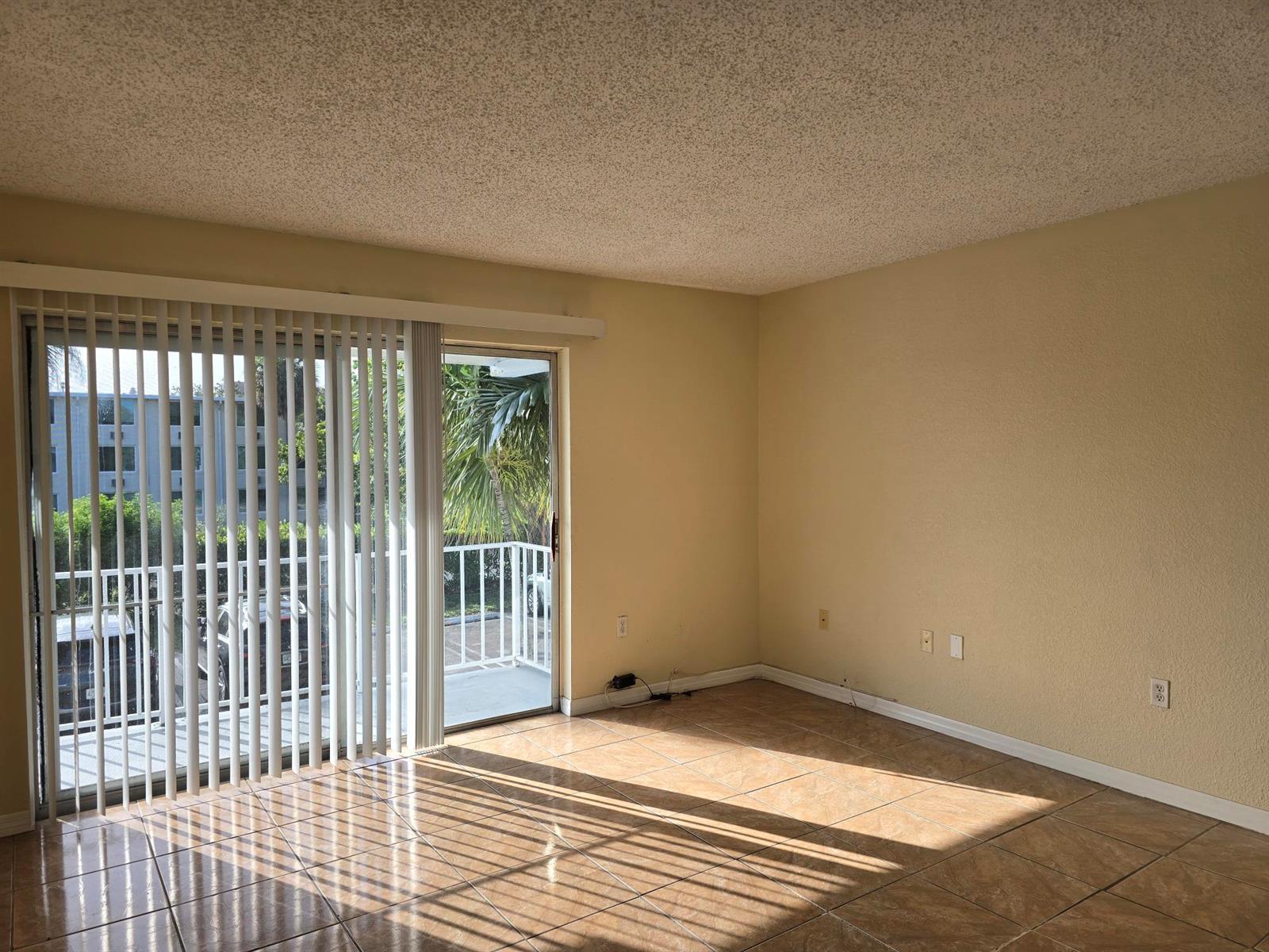 An empty living room with beige walls, tile flooring, and a sliding glass door to a balcony — 411 Executive Center Drive # 210, West Palm Beach FL 33401, WEST PALM BEACH, FL
