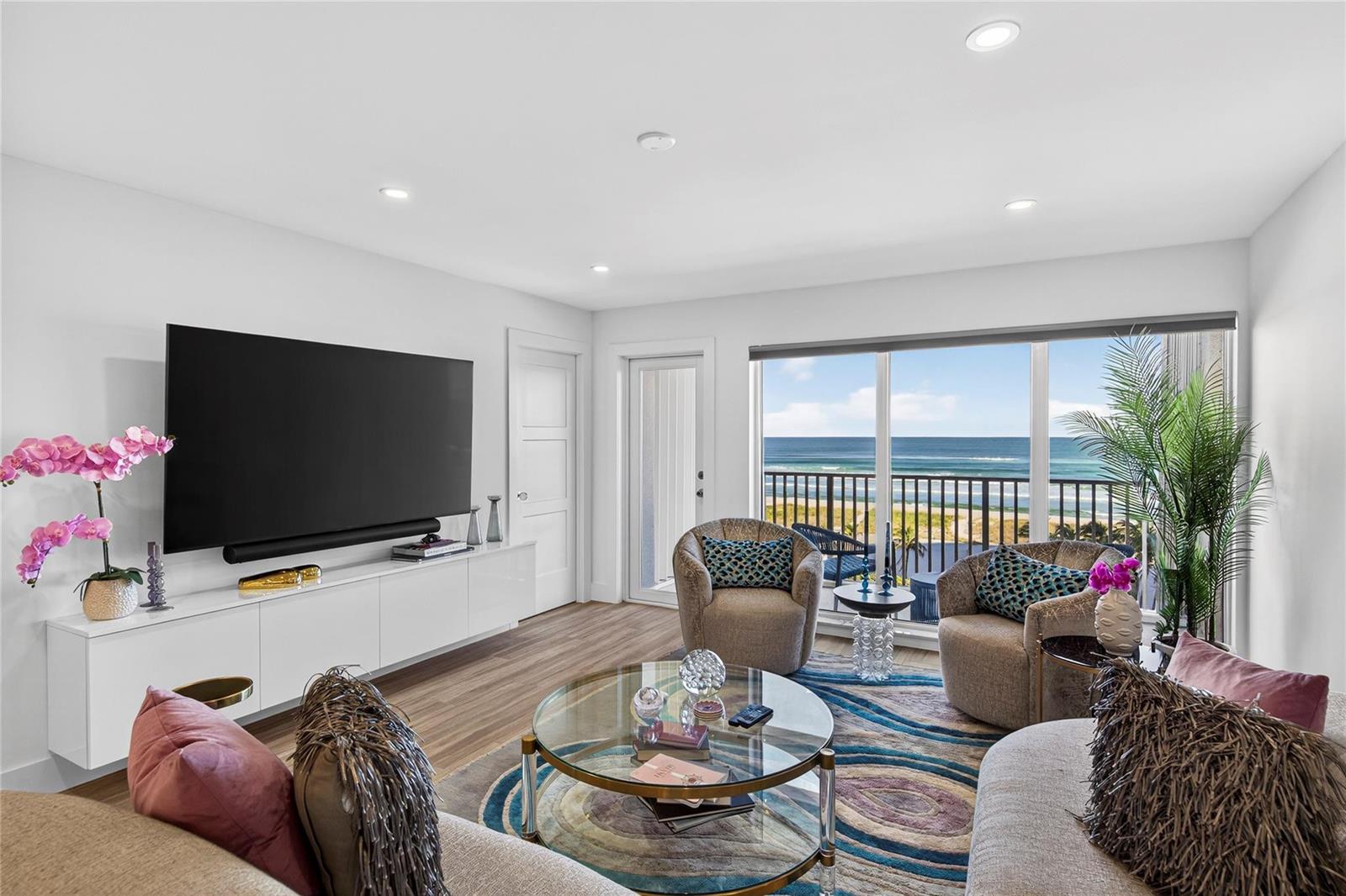 A living room with a large television, circular glass coffee table, and ocean view balcony — 750 N Ocean Boulevard # 608, Pompano Beach FL 33062, POMPANO BEACH, FL