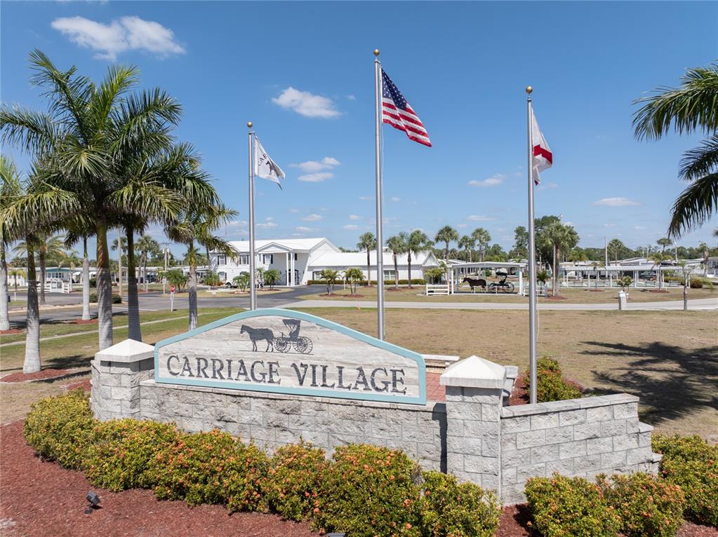 A stone sign for Carriage Village stands in front of a building with three flags — 203 HARVEST LANE, NORTH FORT MYERS, FL