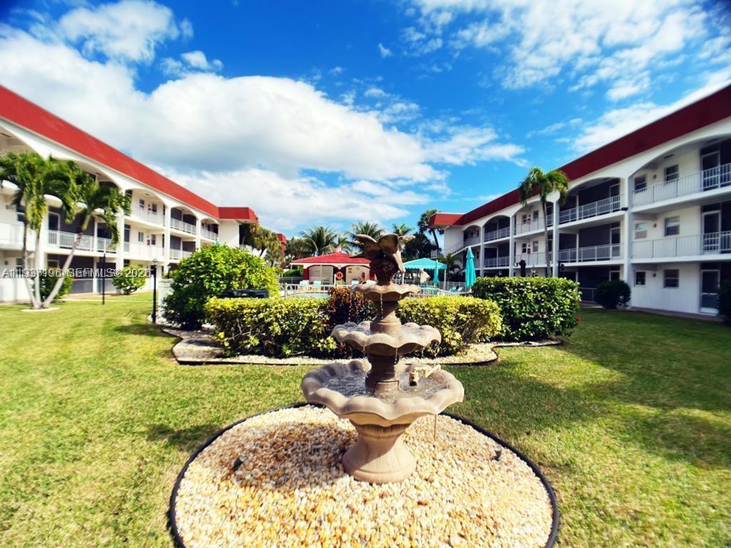 A tiered stone fountain sits in a grassy courtyard between two multi-story apartment buildings — 580 Egret Dr # 302, Hallandale Beach FL 33009, HALLANDALE BEACH, FL