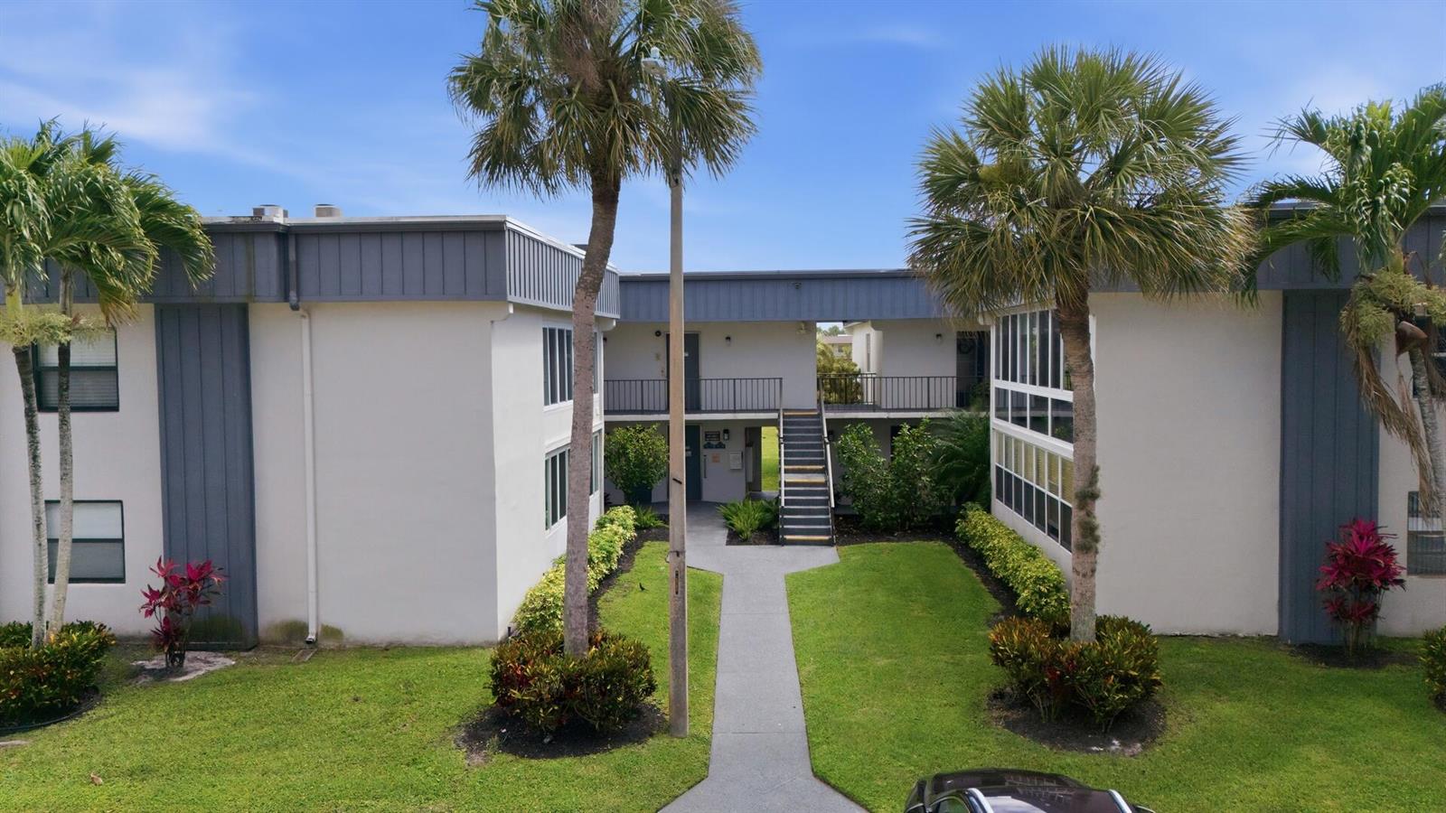 An exterior view of a two-story apartment building with palm trees and a central walkway — 563 Piedmont L, Delray Beach FL 33484, DELRAY BEACH, FL