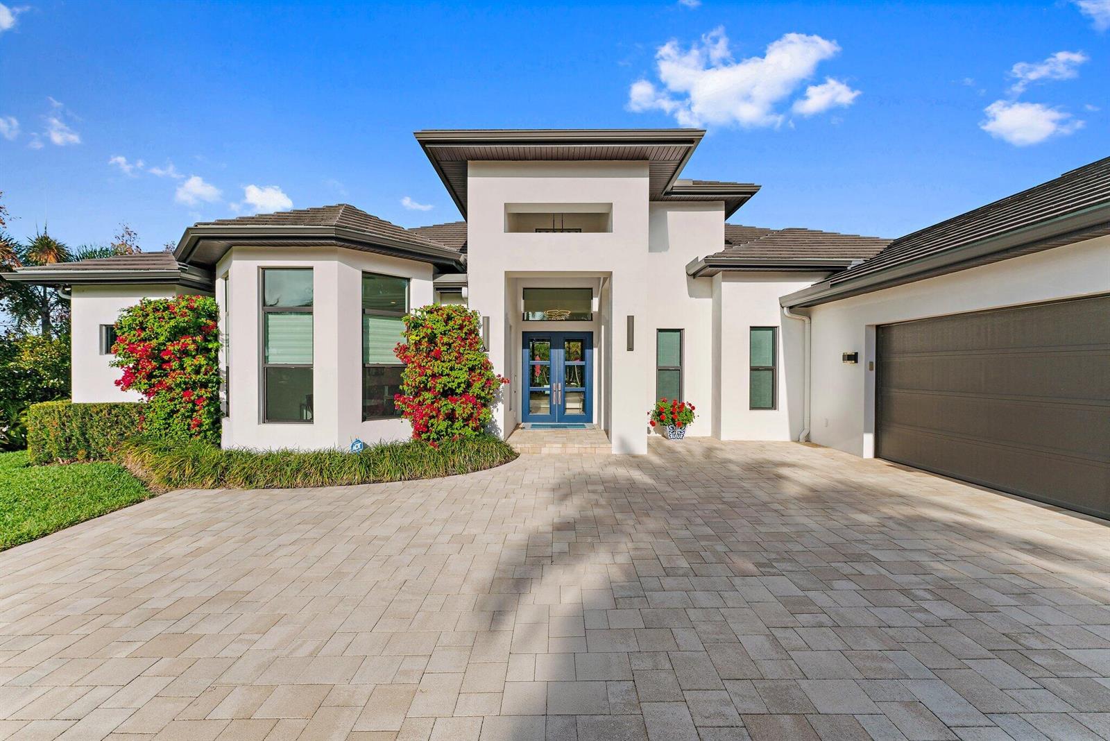 A modern white house features a paved driveway, blue double doors, and red flowering shrubs — 17560 SE Conch Bar Avenue, Jupiter FL 33469, JUPITER, FL