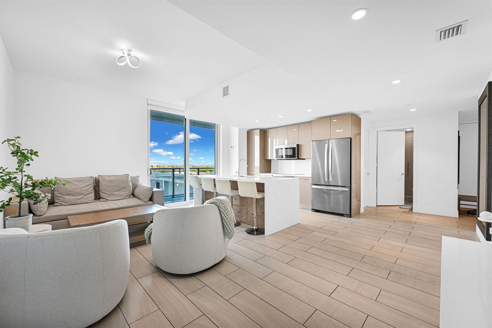 A modern open-concept living room and kitchen with light wood-look tile flooring and balcony access — 2000 Metropica Way # 801, Sunrise FL 33323, SUNRISE, FL