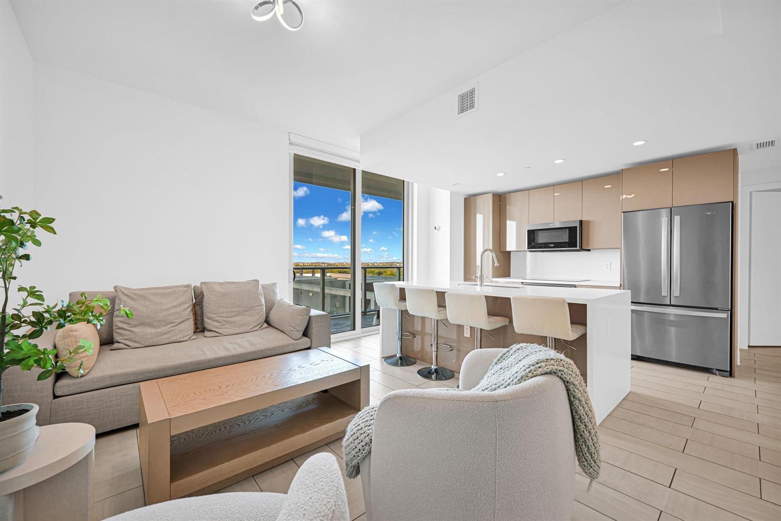 A modern open-concept living room and kitchen with light wood-toned cabinets and a balcony view — 2000 Metropica Way # 801, Sunrise FL 33323, SUNRISE, FL