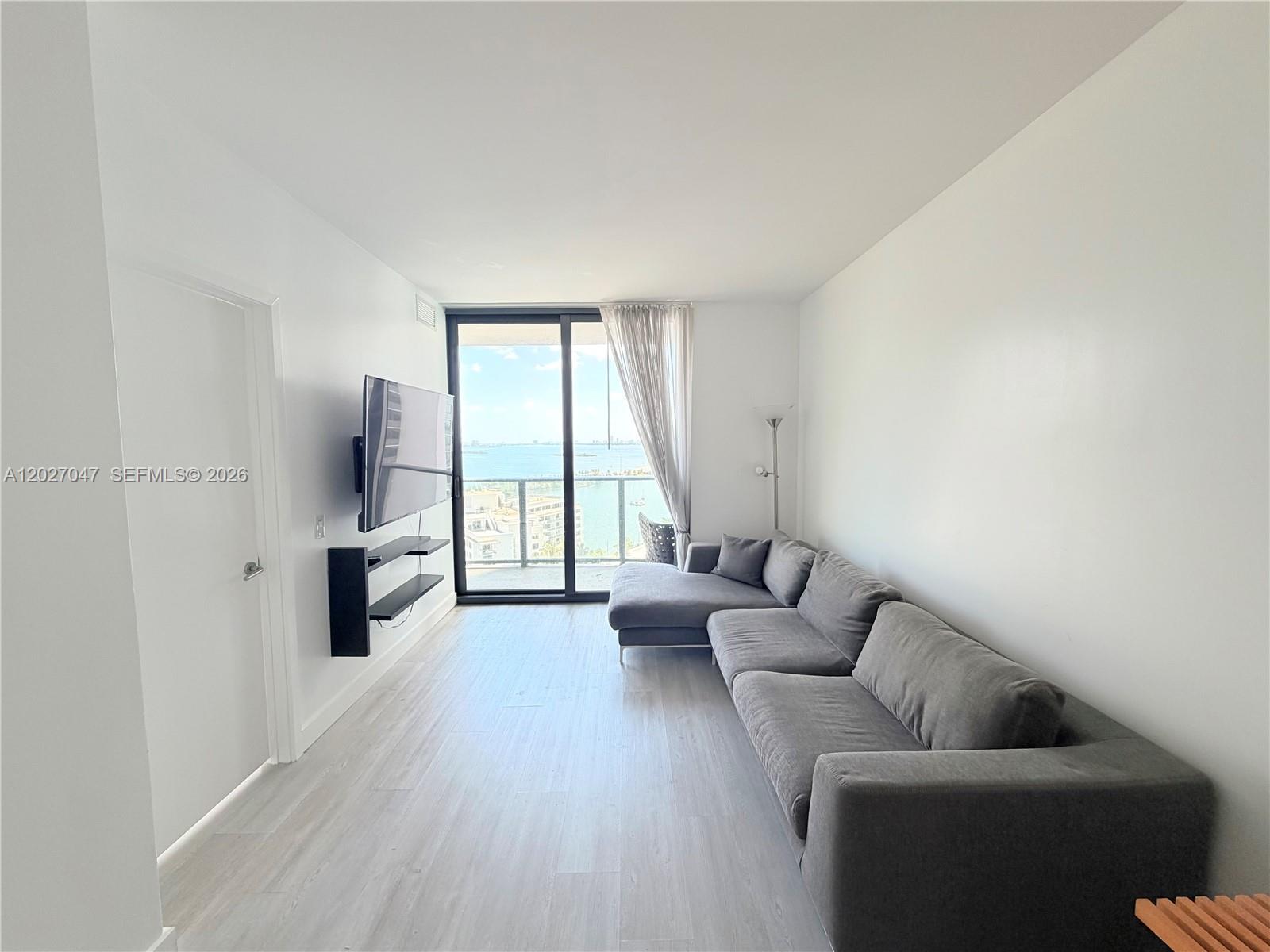 A living room features a grey sectional sofa, wall-mounted television, and a balcony with water views — 501 NE 31st St # 1804, Miami FL 33137, MIAMI, FL