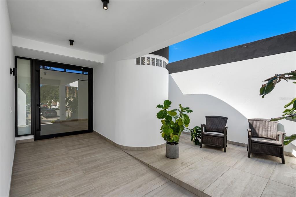 A modern covered patio features light wood-look tile flooring, white curved walls, and two wicker chairs — 3 GARDEN HILLS ESTATES UNIT 12, GUAYNABO, PR