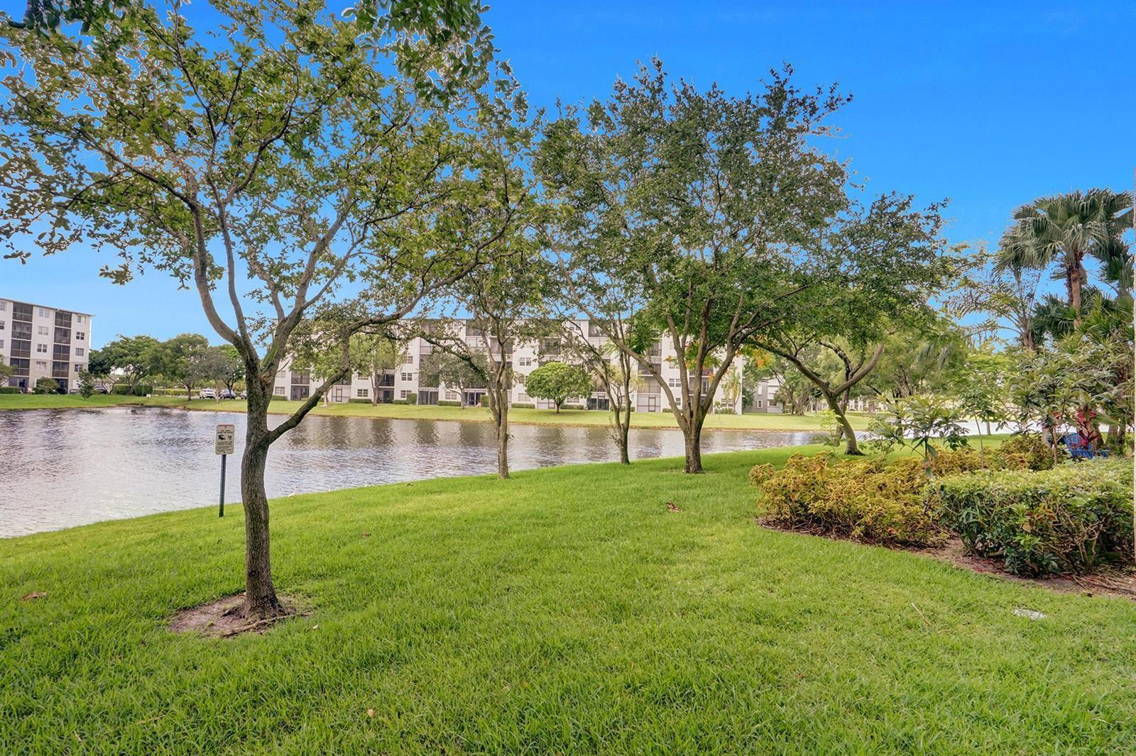 A grassy lawn with trees overlooks a lake in front of a multi-story apartment building — 2334 S Cypress Bend Drive # 907, Pompano Beach FL 33069, POMPANO BEACH, FL