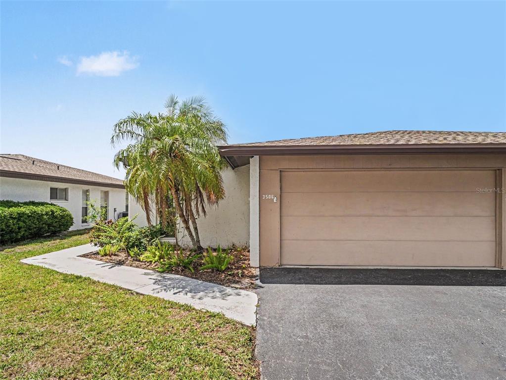 A tan garage door is attached to a white stucco house with a palm tree — 3506 AUBURN COURT UNIT 230, SARASOTA, FL