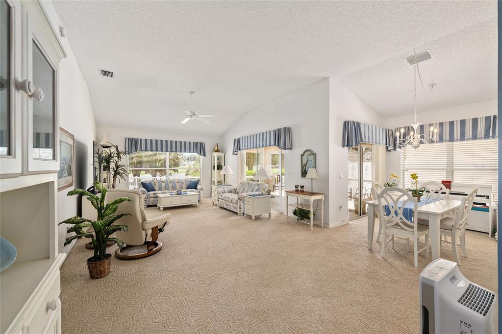 A spacious living and dining area with vaulted ceilings, beige carpeting, and blue-striped window valances — 17239 SE 84TH KNIGHT AVENUE, THE VILLAGES, FL