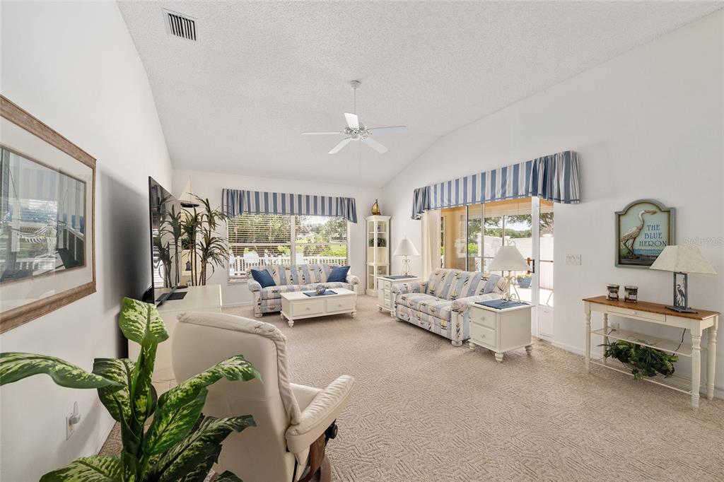 A living room features a vaulted ceiling, beige carpeting, two sofas, and a sliding glass door — 17239 SE 84TH KNIGHT AVENUE, THE VILLAGES, FL