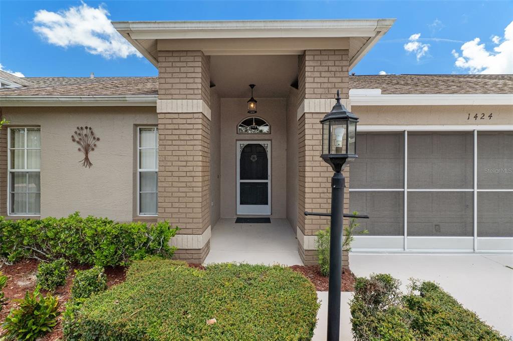 A tan house exterior features a brick-accented entryway, a screen door, and a lamp post — 1424 PINEWOOD HILLS COURT, SPRING HILL, FL
