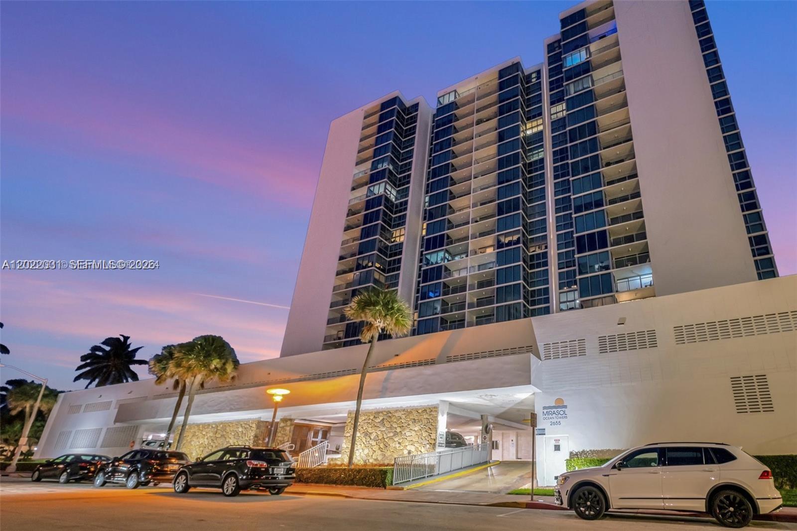 A tall residential building with a stone-accented entrance and parking area at sunset — 2655 Collins Ave # 2302, Miami Beach FL 33140, MIAMI BEACH, FL