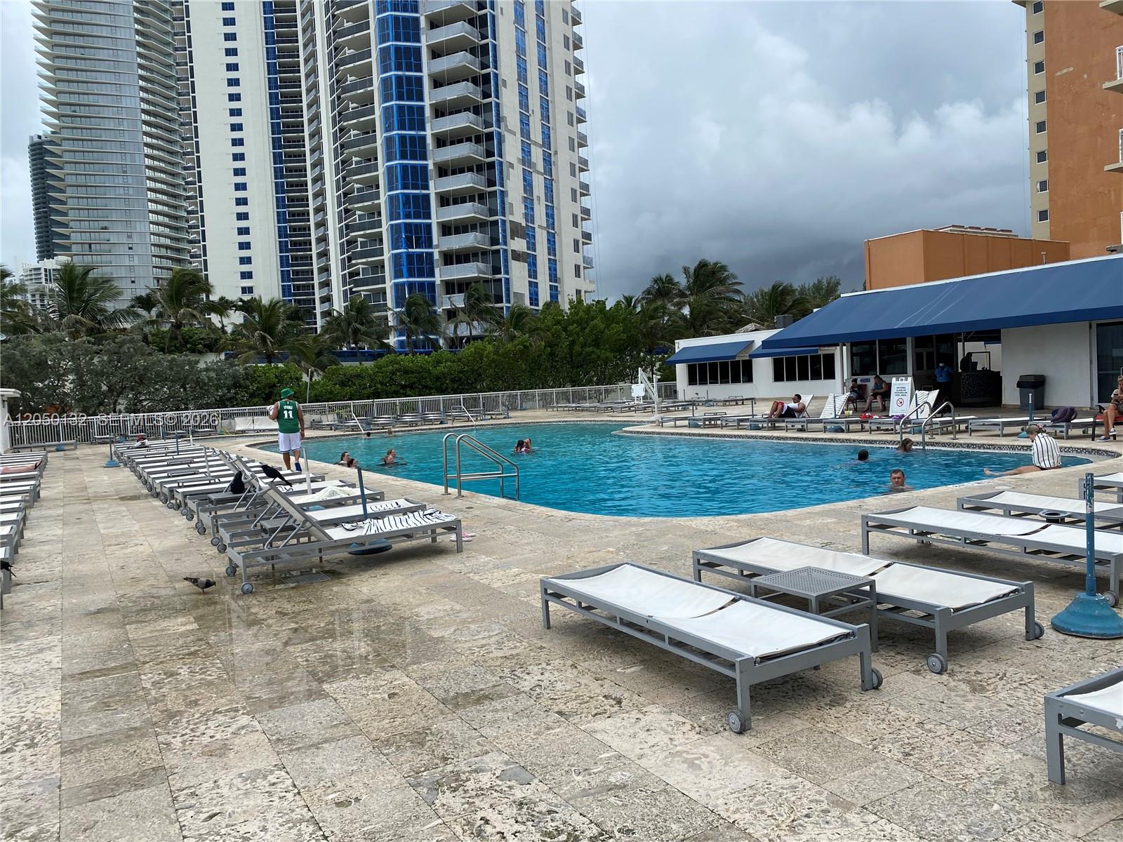 An outdoor swimming pool area features lounge chairs on a stone deck near tall buildings — 19201 E Collins Ave # 442, Sunny Isles Beach FL 33160, SUNNY ISLES BEACH, FL