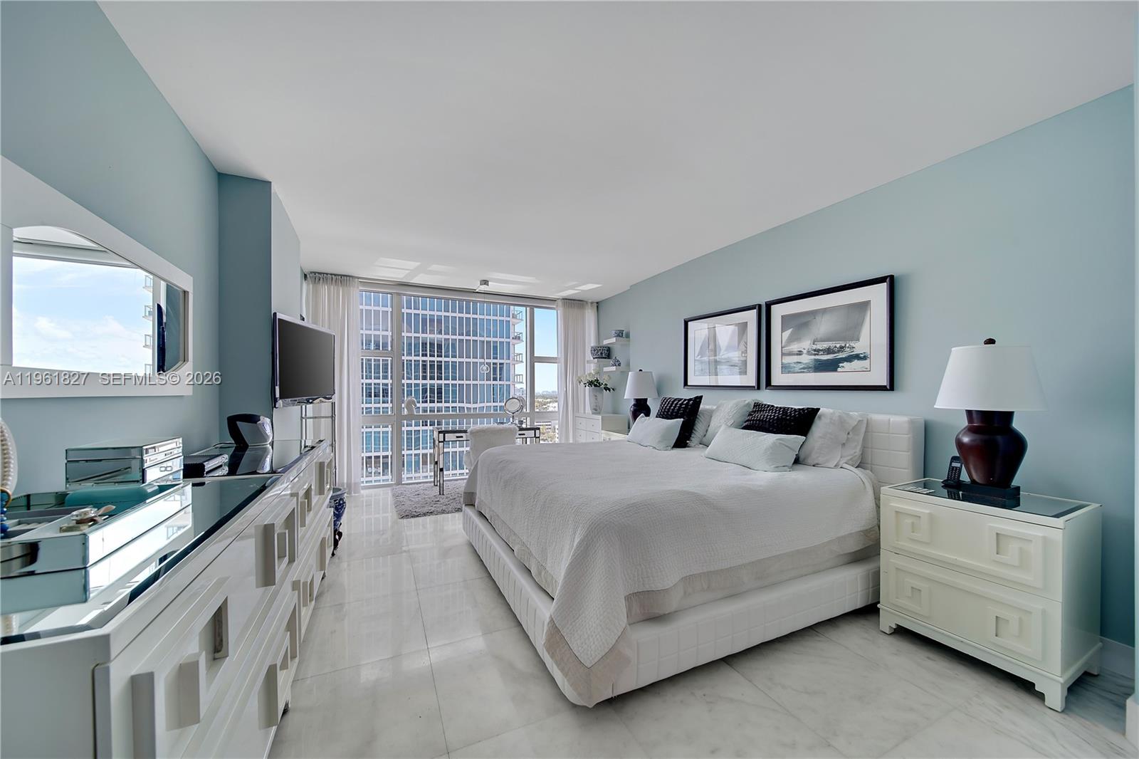 A bright bedroom features a white bed, mirrored furniture, and floor-to-ceiling windows overlooking a city — 6801 Collins Ave, Miami Beach FL 33141, MIAMI BEACH, FL