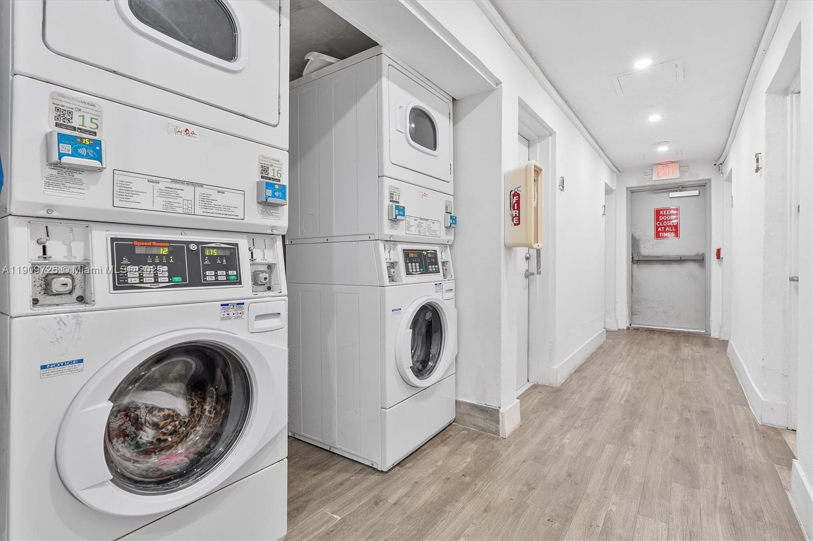 A laundry room features two stacked washer-dryer units, light wood-look flooring, and a fire exit — 420 78th St, Miami Beach FL 33141, MIAMI BEACH, FL