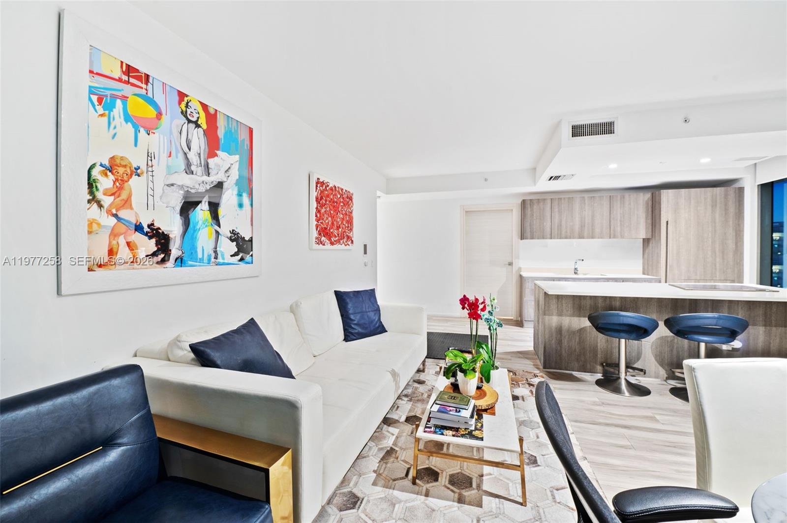 A modern living room features a white sofa, blue accents, and an open-concept kitchen area — 801 S Miami Ave # 2705, Miami FL 33130, MIAMI, FL