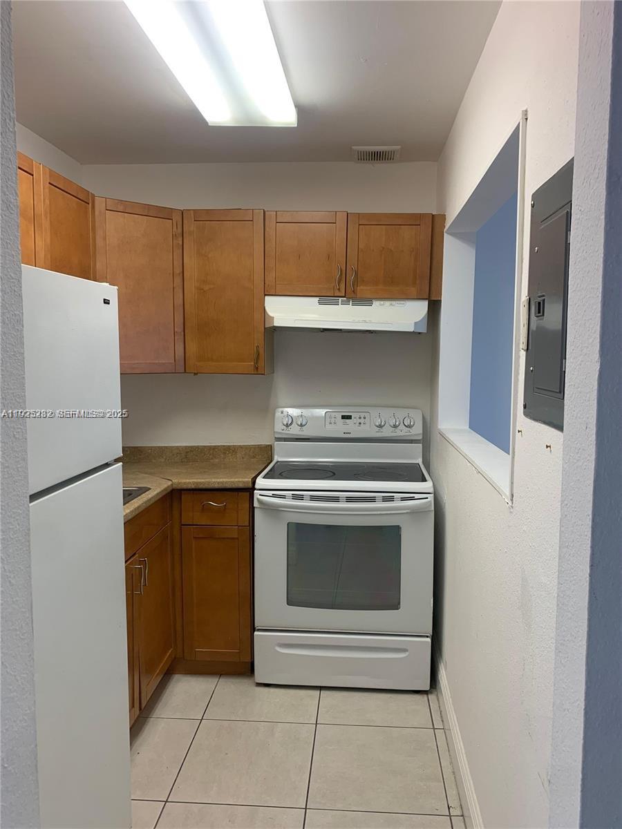 A small kitchen features wooden cabinets, white appliances, and light-colored floor tiles — 241 SE 9th Ave # 106, Pompano Beach FL 33060, POMPANO BEACH, FL
