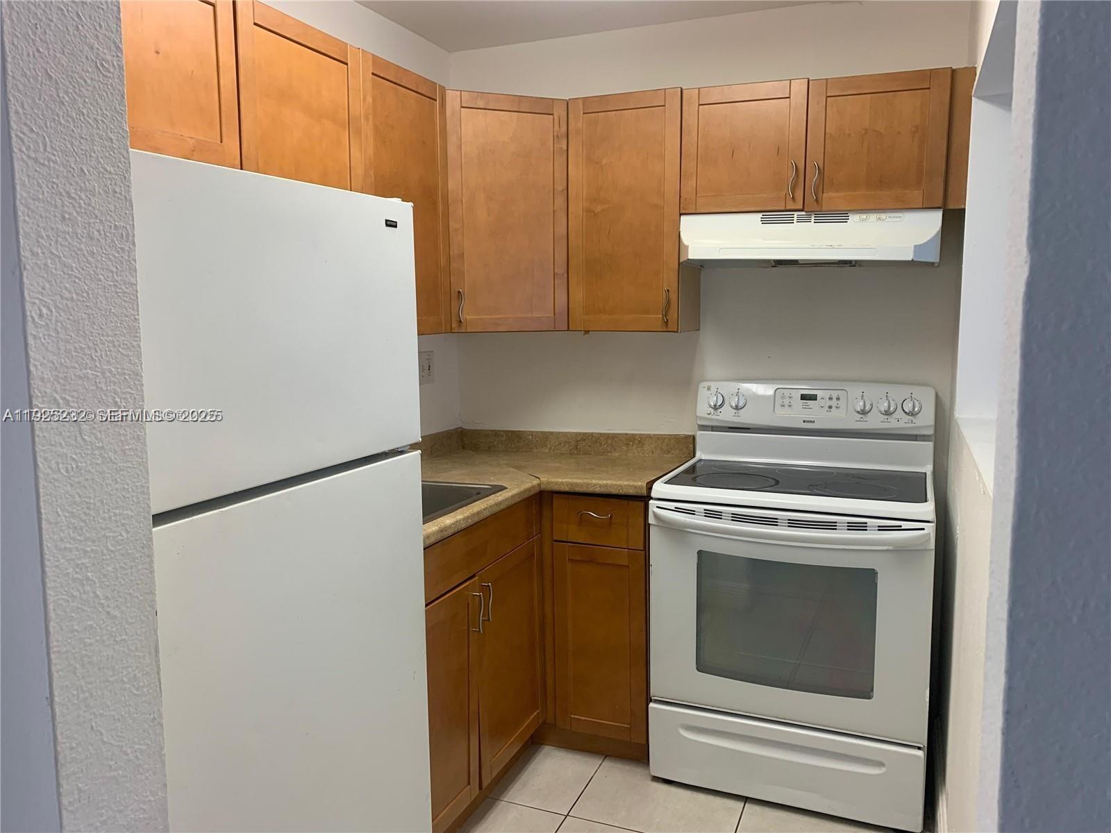 A small kitchen features wooden cabinets, a white refrigerator, a white stove, and countertops — 241 SE 9th Ave # 106, Pompano Beach FL 33060, POMPANO BEACH, FL